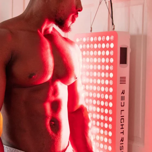 A toned, muscular man stood in front of a red light therapy panel hanging on a door